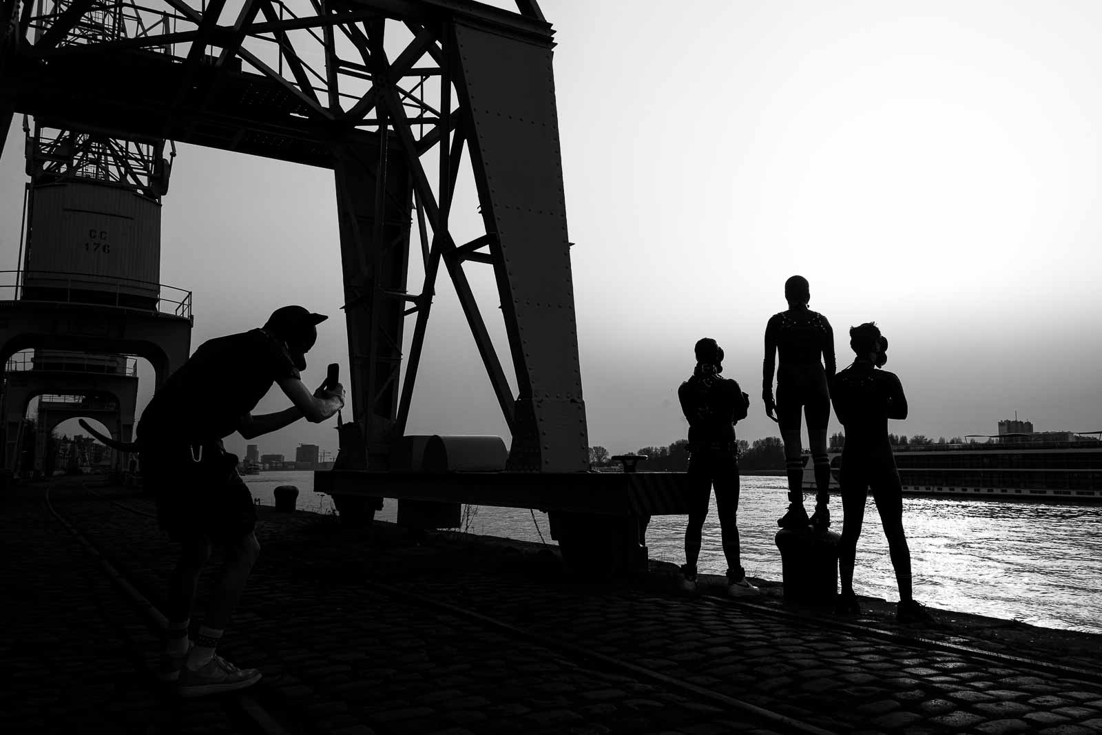 A black and white silhouette photograph of a photographer taking a picture of three individuals posing near a large industrial crane next to a river.