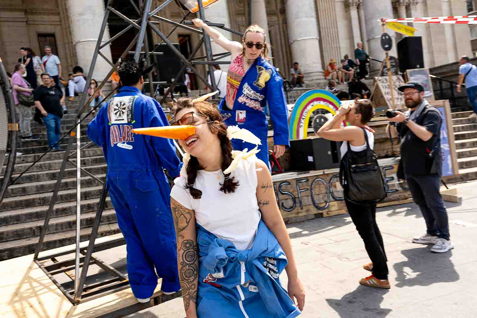 A performer wearing a blue jumpsuit and a long fake carrot nose laughs joyfully in the foreground, with other performers climbing a metal structure behind them at the Zinneke parade.